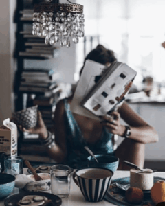 A stylishly dressed woman having a leisurely, intentional breakfast under a crystal chandelier while reading a magazine or book, symbolizing the design of a focused morning routine.