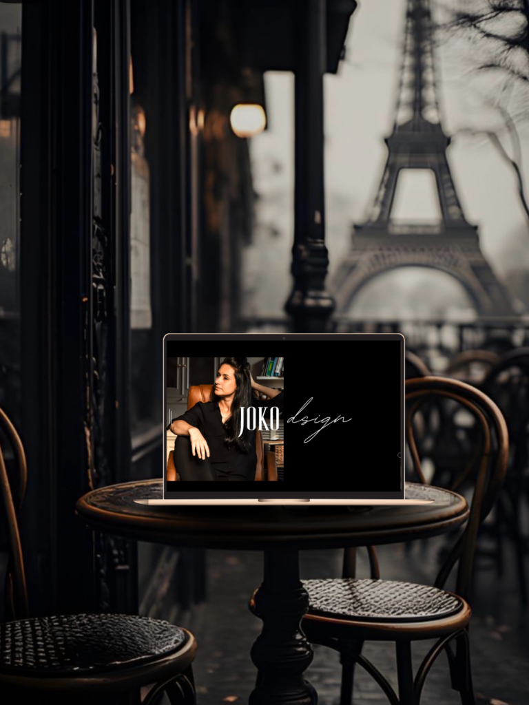 A gold laptop mockup sitting on a round bistro table in a dark, moody Parisian cafe. The screen displays a luxury website design for "JOKO design" with a dark background and elegant script typography. The Eiffel Tower is visible in a soft, blurred background.